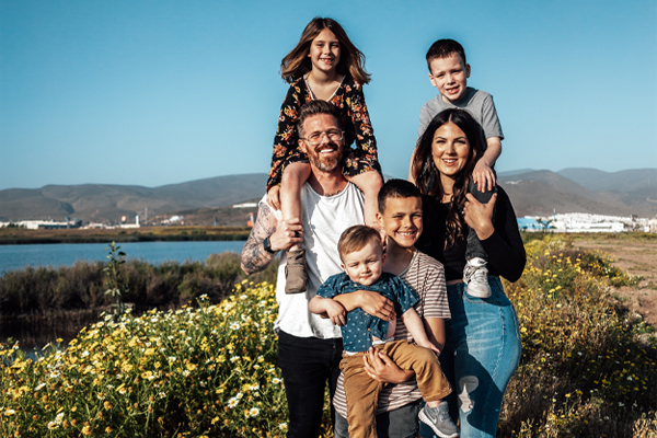 Big happy family standing together in front of a lake. Husband holding daughter on his shoulders, wive holding son on hers, and another two sons standing in front of them one holding the other.